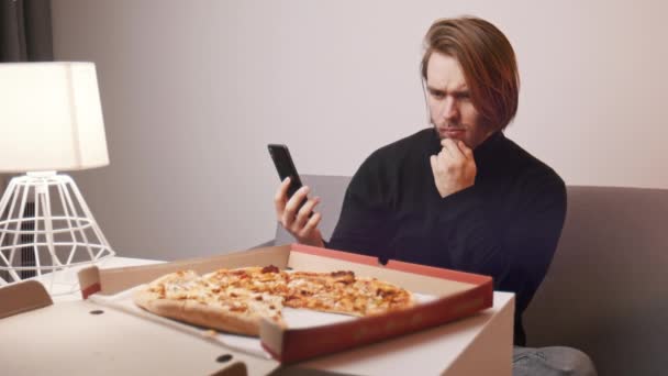 En ung kaukasisk man som klagar på pizza. Hålla smartphone för att ringa kundsupport — Stockvideo