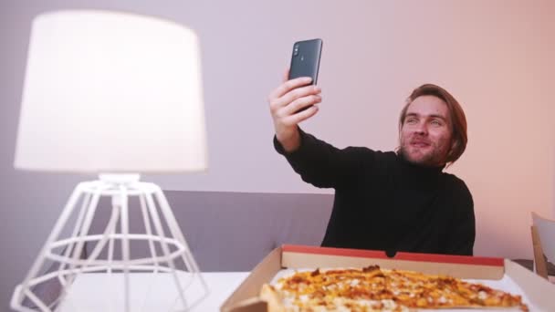 Młody biały człowiek mając wideo rozmowy na jego smartfon podczas jedzenia pizzy na obiad — Wideo stockowe
