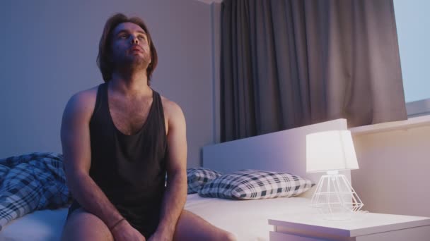 Молодой кавказский мужчина сидит на кровати. Бессонница, психическое расстройство или депрессия — стоковое видео