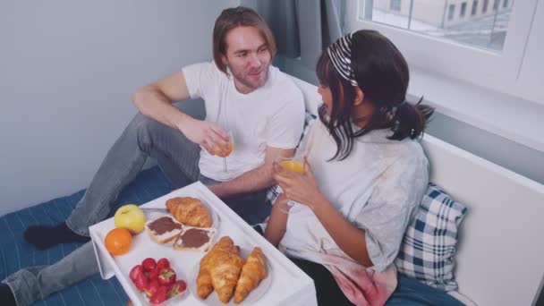 Ungt flerkulturelt par som drikker appelsinjuice og spiser frokost på sengen. Vakker flerrasefamilie – stockvideo