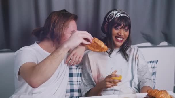 Jong gelukkig stel dat plezier heeft tijdens het ontbijt op bed. Man geeft croissant aan zijn vrouw — Stockvideo