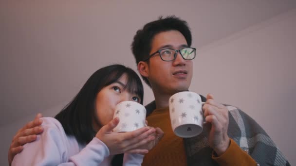 Jong pasgetrouwd Aziatisch koppel dat warme drank drinkt en naar een film kijkt bedekt met deken. Liefde en gezinsconcept — Stockvideo