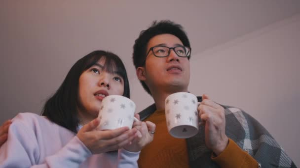 Jovem família feliz, casal asiático bebendo bebida quente e assistindo TV — Vídeo de Stock