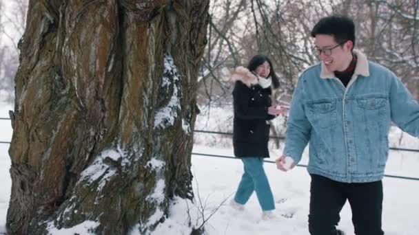 Snö spel. Glada unga asiatiska par kastar snöbollar och springer runt trädet — Stockvideo