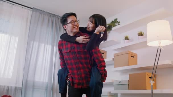 Mladý asijský pár se baví v novém bytě. Přítel dává piggyback jízda na jeho dívčí fliend, zatímco ona drží klíče — Stock video