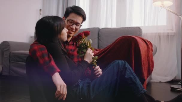 Joven pareja asiática pasando aniversario en el nuevo apartamento. Mujer sosteniendo ramo de rosas rojas. Ver la televisión — Vídeo de stock