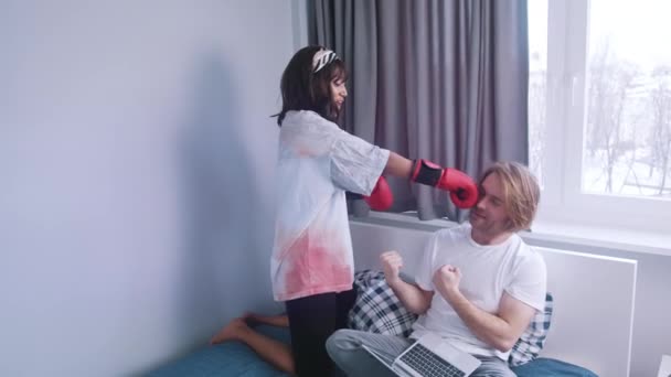 Dziewczyna bijąca chłopaka rękawiczkami bokserskimi. Kobieta szuka uwagi. Człowiek pracujący na laptopie w łóżku — Wideo stockowe