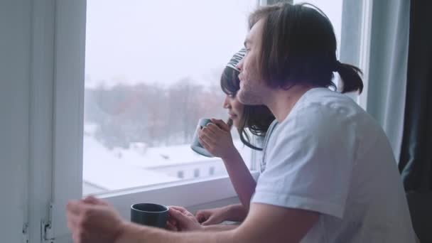 Gelukkig multiraciaal paar drinken warm drankje terwijl kijken door het raam op het eerste sneeuw — Stockvideo