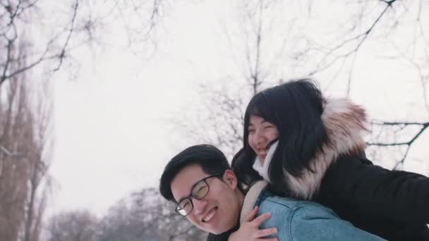 Young man giving piggyback ride to his girlfriend and spinning her in the park on snowy winter day. Newlywed asian couple having fun outside — Stock Video