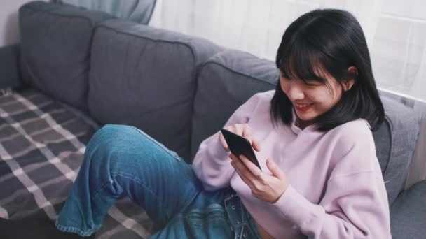 Jeune femme asiatique se détendre sur le canapé et sourire tout en lisant le message sur le smartphone — Video