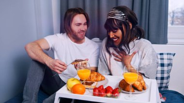 Genç, çok ırklı, yeni evli çift balayında yatakta kahvaltı yapıyor.