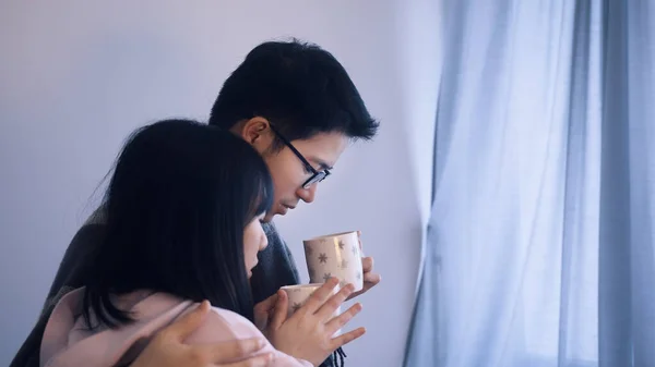 Casal jovem bebendo bebida quente e olhando pela janela coberta com cobertor. Conceito férias de inverno — Fotografia de Stock