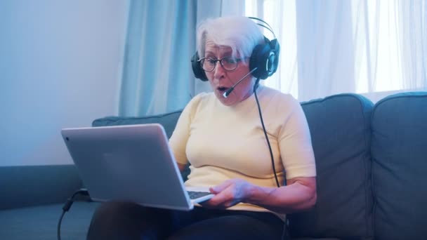 Překvapená starší žena se sluchátky mávajícími před kamerou na notebook. Uzamčení a zábava — Stock video