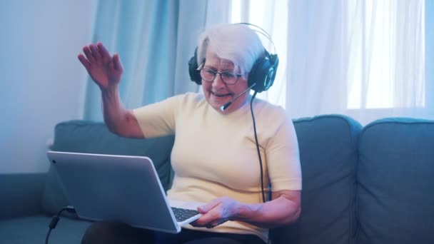Starší žena se sluchátky, která má videohovor s rodinou a mává před kamerou. Staří lidé a technologie — Stock video