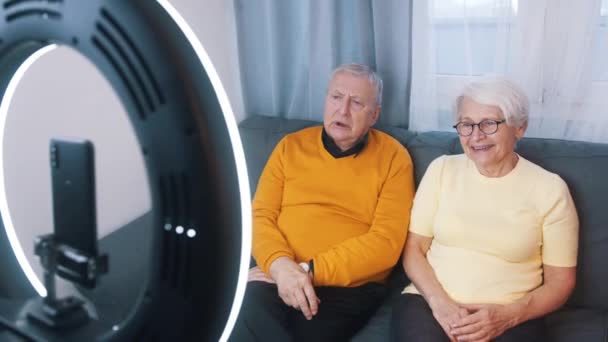 Пожилая пара дает интервью или записывает видео сообщение для семьи. Женщина машет на камеру смартфона — стоковое видео