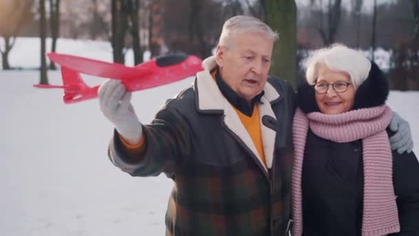 Glückliches älteres Paar, das sich im Winter im Park umarmt und mit Flugzeugspielzeug spielt. Zeitlupe — Stockvideo