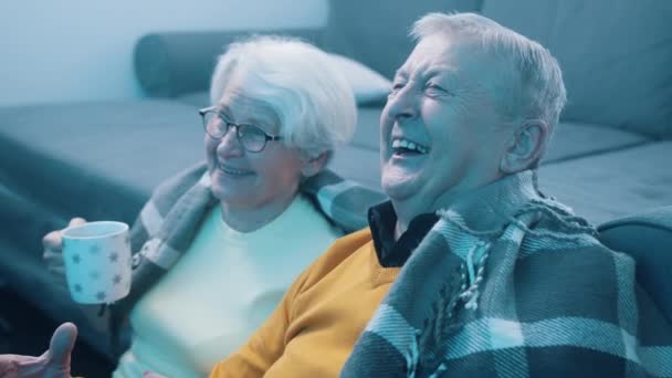 Pasangan tua yang bahagia tertawa menonton TV dan minum minuman panas yang ditutupi dengan selimut — Stok Video