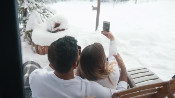 Unga par sitter på bänken utomhus och dricker varm choklad och tar selfie på snöiga Alla hjärtans dag — Stockvideo