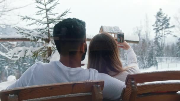 Vacances d'hiver. Couple multiracial assis sur le banc en plein air Femme prenant selfie sur la Saint-Valentin enneigée — Video