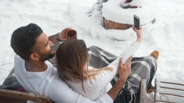Multiraciaal paar zitten op de bank buiten het drinken van warme chocolademelk en het nemen van selfie op besneeuwde Valentijnsdag — Stockvideo