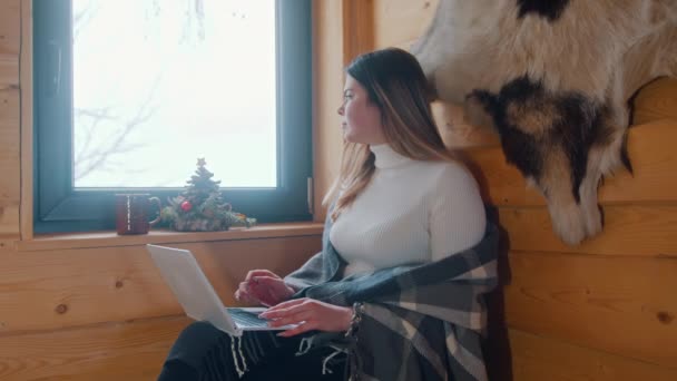 Lonely young caucasian woman sitting near the window covered with blanket lookin outside and using laptop — Stock Video