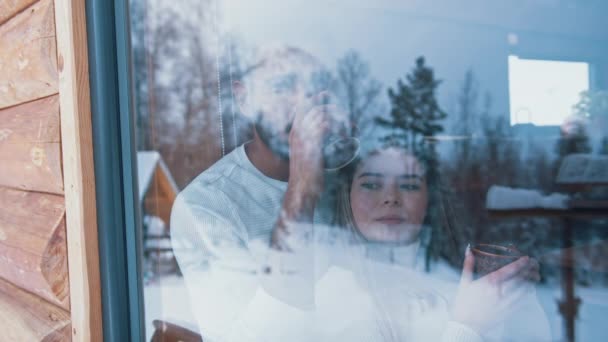 Ευτυχισμένο ζευγάρι που πίνει ζεστό ρόφημα αγκαλιάζοντας και κοιτώντας από το παράθυρο. Χειμερινές διακοπές, Ημέρα του Αγίου Βαλεντίνου ή Χριστούγεννα — Αρχείο Βίντεο