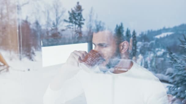 Junger Mann trinkt Heißgetränk und blickt durch das Fenster auf den ersten Schnee. Weihnachtsfeier — Stockvideo