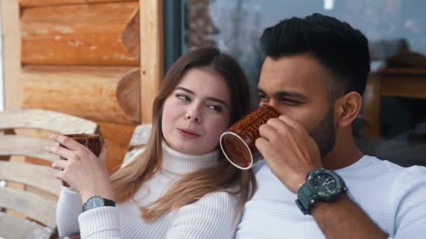 De cerca. Feliz joven pareja abrazándose cubierta con manta en el banco al aire libre. Beber bebidas calientes y disfrutar de la nieve — Vídeos de Stock