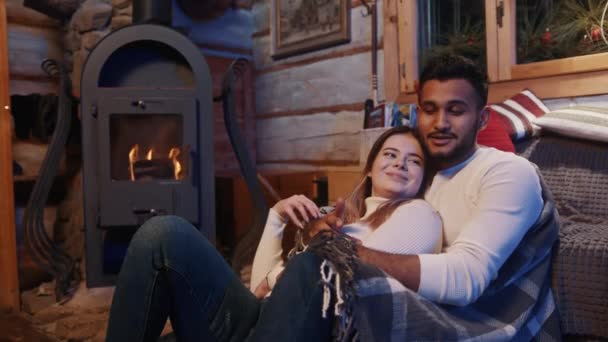 Feliz casal multirracial jovem relaxante perto da lareira coberta com cobertor. Passar férias de inverno juntos — Vídeo de Stock