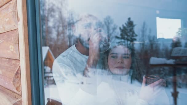 Feliz joven pareja bebiendo bebida caliente abrazándose y mirando por la ventana. Vacaciones de invierno, San Valentín o Navidad — Vídeos de Stock