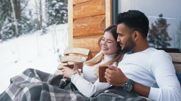 Feliz joven pareja abrazándose cubierta con manta en el banco al aire libre. Beber bebidas calientes y disfrutar de la nieve — Vídeos de Stock