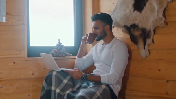 Молодой индиец сидит у окна в уютном доме и работает на ноутбуке, выпивая горячий напиток — стоковое видео