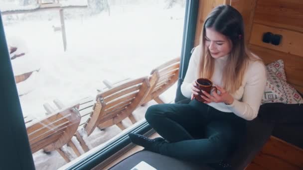 Mujer caucásica joven bebiendo café y utilizando el ordenador portátil cerca de la ventana. Vacaciones de invierno — Vídeo de stock