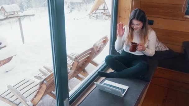 Βίντεο κλήση. Νεαρή γυναίκα χαιρετάει την κάμερα του laptop πίνοντας καφέ κοντά στο μεγάλο παράθυρο στις χειμερινές διακοπές — Αρχείο Βίντεο