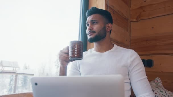 ハンサムなインド人男性飲料コーヒーの肖像画と大きな窓の近くのラップトップで作業 — ストック動画