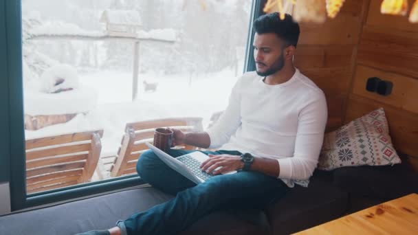 Vacaciones de invierno. Joven navegando por las redes sociales y bebiendo té cerca de la ventana — Vídeo de stock