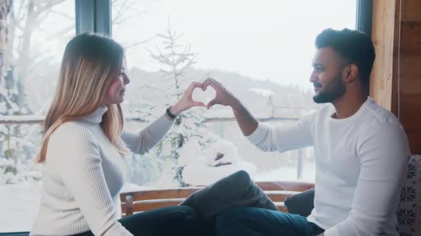 Feliz Dia dos Namorados. Jovem casal multirracial fazendo forma de coração com as mãos perto da janela. Neve lá fora — Vídeo de Stock