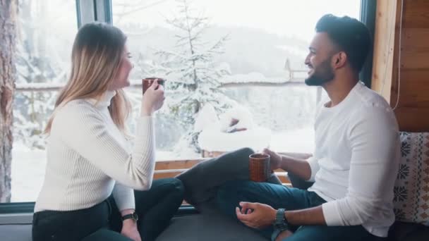 Feliz pareja multirracial bebiendo bebidas calientes y mirando la nieve afuera. Vacaciones de invierno, Navidad o San Valentín — Vídeos de Stock