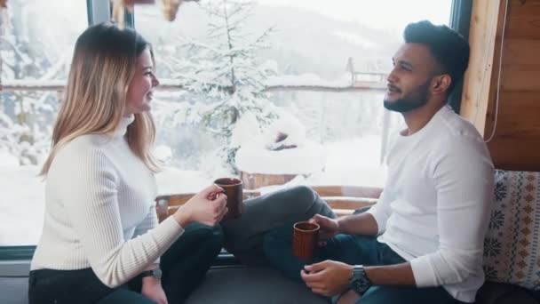 Joyeux jeune couple qui boit une boisson chaude et regarde la neige dehors. Vacances d'hiver, Noël ou Saint Valentin — Video