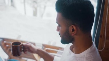 Genç Hintli adam dizüstü bilgisayarda çalışıyor ve karla kaplı ağaçlara bakarken sıcak çikolata içiyor.
