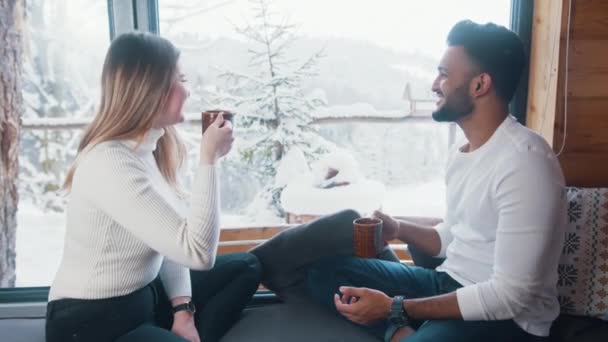 Молода багаторасова пара насолоджується зимовими канікулами. Дивлячись на сніг через вікно і пити чай — стокове відео