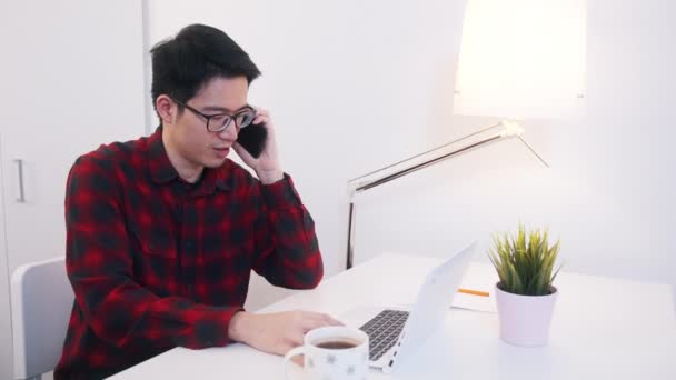 Jovem asiático homem falando no telefone enquanto workingo n laptop no escritório — Vídeo de Stock
