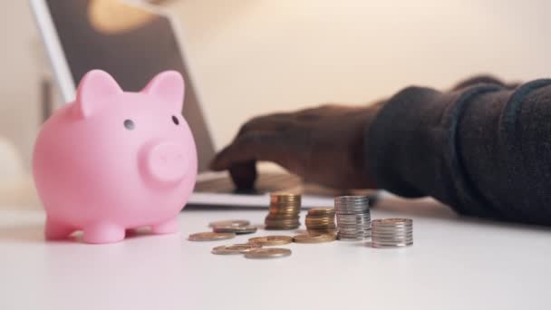 Manos de hombre negro trabajando en el portátil detrás de la hucha y las monedas. Inversiones y gestión del concepto de riesgo — Vídeo de stock