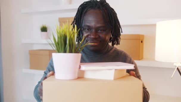 Возбужденный молодой африканский американец переезжает в новую квартиру. Держать картонные коробки с растениями и книгами — стоковое видео