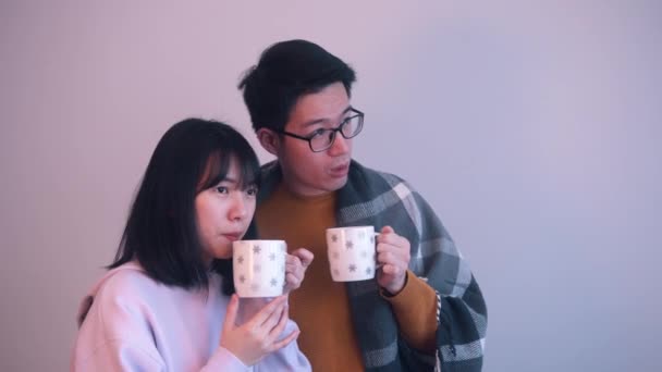Молодая азиатская пара обнимается под одеялом и пьет горячий напиток в закрытом помещении в бурный день — стоковое видео