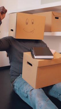 Dikey, mutlu, kafasında karton kutuyla genç bir adam yeni dairede el dansı yapıyor.