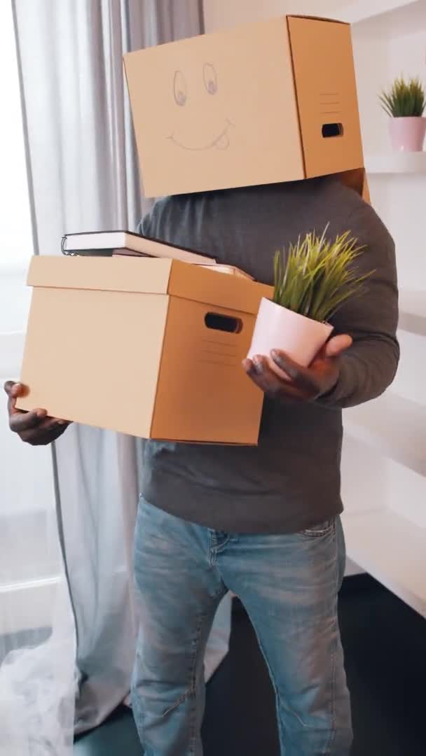 Vertical, méconnaissable homme afro-américain avec boîte en carton sur la tête et plante à la main se déplaçant dans un nouvel appartement — Video