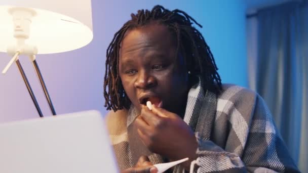 Hombre negro enfermo con fiebre envuelto en blanco tomando una píldora mientras recibe consultas médicas remotas — Vídeo de stock