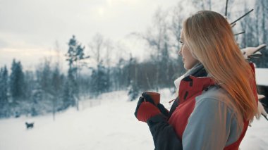Genç sarışın kadın sıcak bir içecek içiyor ve dağda kar yağışı seyrediyor.