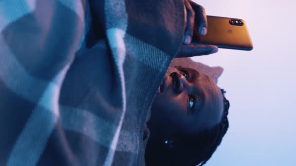 Vertikaler, junger schwarzer Mann checkt die sozialen Medien auf seinem Handy, bedeckt mit einer Decke auf der Couch — Stockvideo